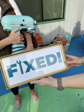 Woman holding a KitchenAid stand mixer at a Repair Fair with a "Fixed" sign in front of it.