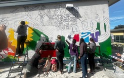Teens work on a mural on the side of a school building.