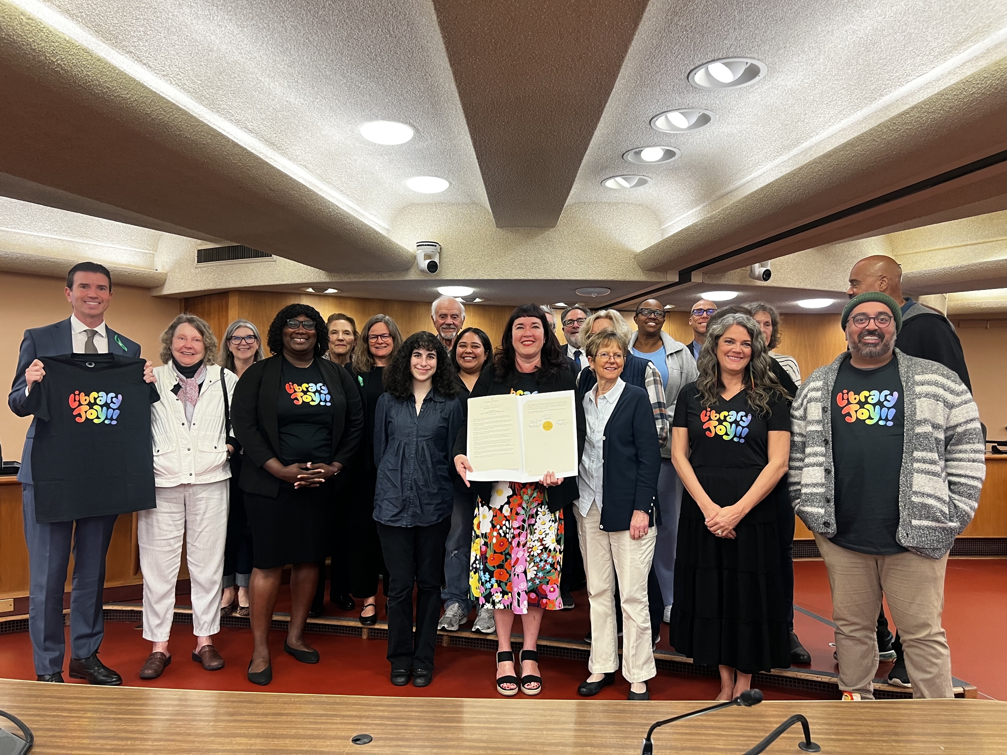 Board, Library staff, and Friends of the Library smiling holding resolution in Board chambers