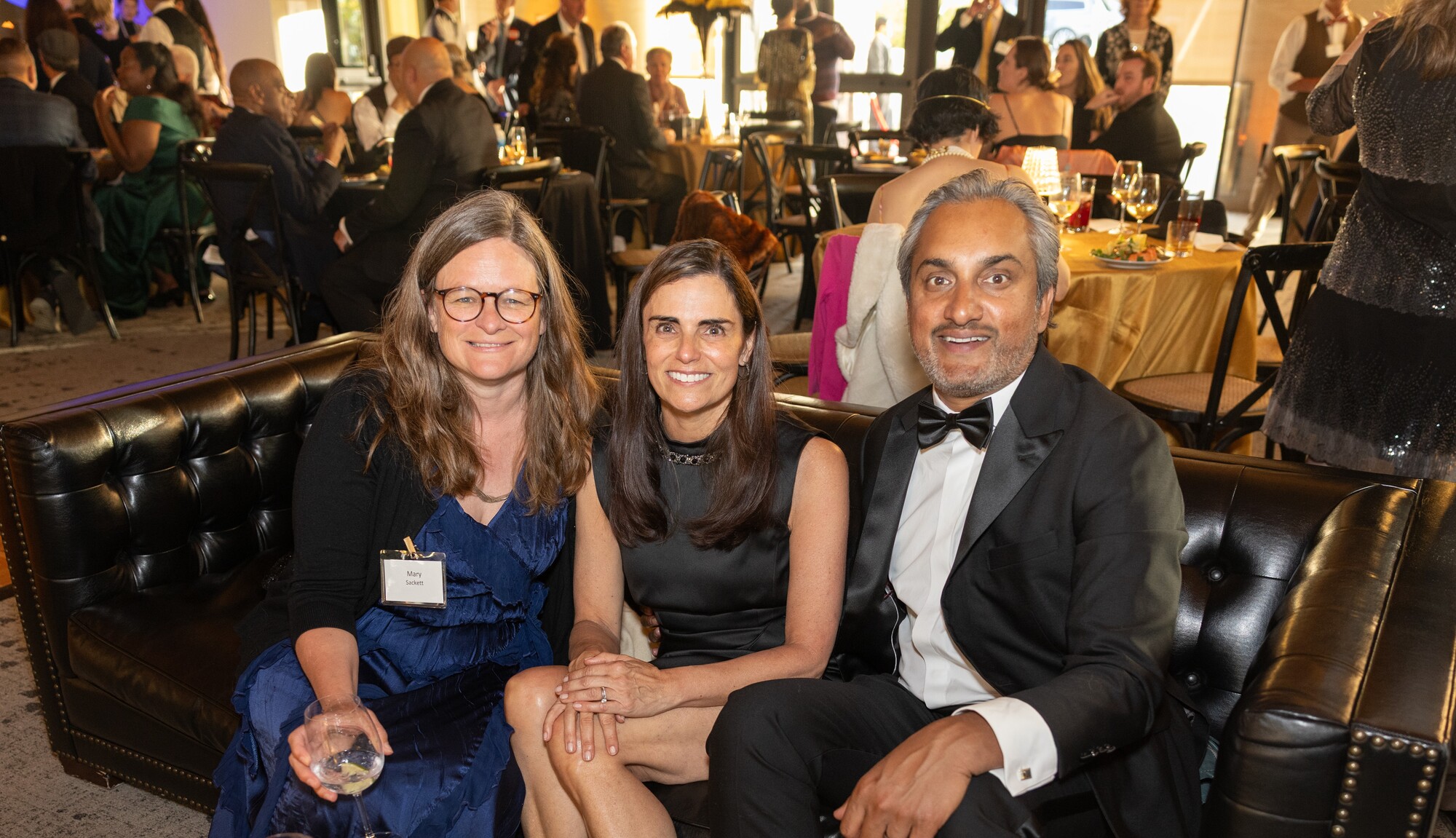 Mary, Heather, Sumant smiling at gala seated on couch with crowd in background