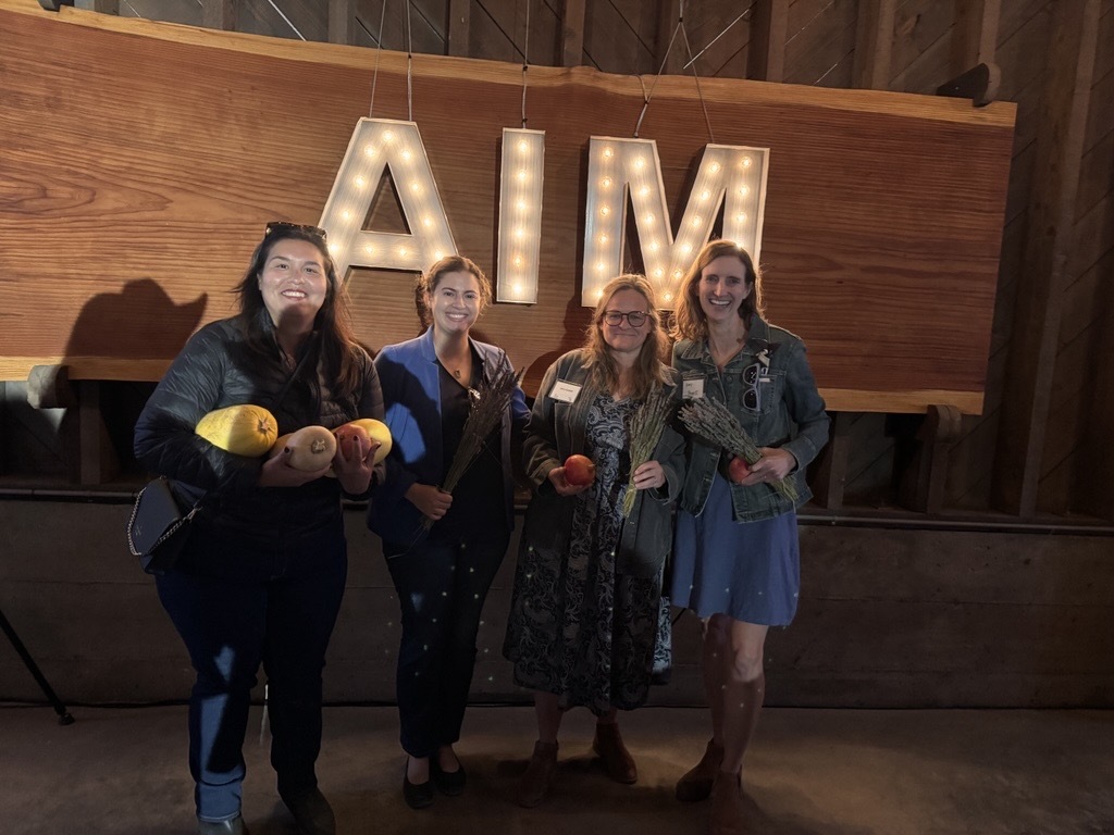 Mary, Crystal, Julia, and friend smiling holding lavender bundles and fall produce under light up letters spelling AIM