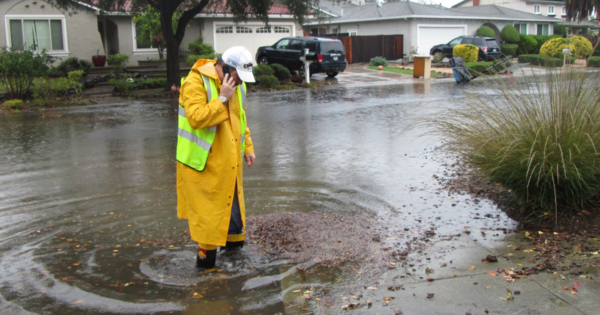 Man in yellow rain jacket and neon vest on phone standing in the middle of a flooded street