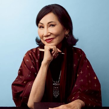 Amy Tan in burgundy shall holding a pen in right hand and smiling into the camera