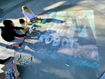 Photo of children using chalk to create the book cover for The Wild Robot on the sidewalk outside the Library