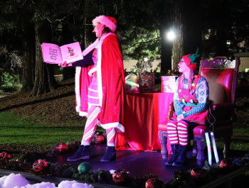 Photo of Mrs. Claus reading a book while the Library Elf watches on during the Tree Lighting Celebration