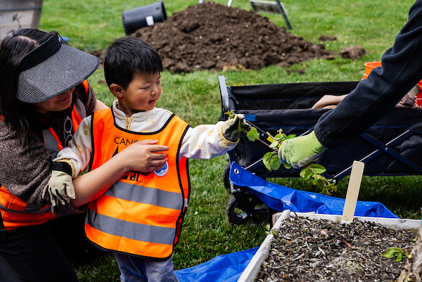 Picture of a child in a bright orange vest reaching out to touch a plant being offered by an adult.