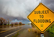 Photo of a yellow warning sign that reads "subject to flooding" nest to a flooded roadway. 