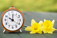 Photo of an analog clock on a table next to flowers. 