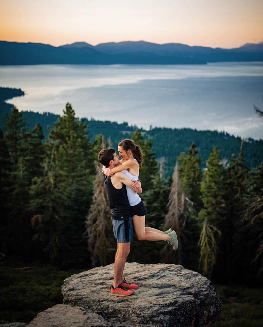 Two people hugging on a rocky hill top in a forest with lake in the background during sunset