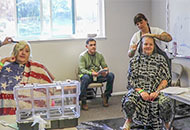 two people sitting in barbers chairs getting their haircut by barbers as a man waits in the background