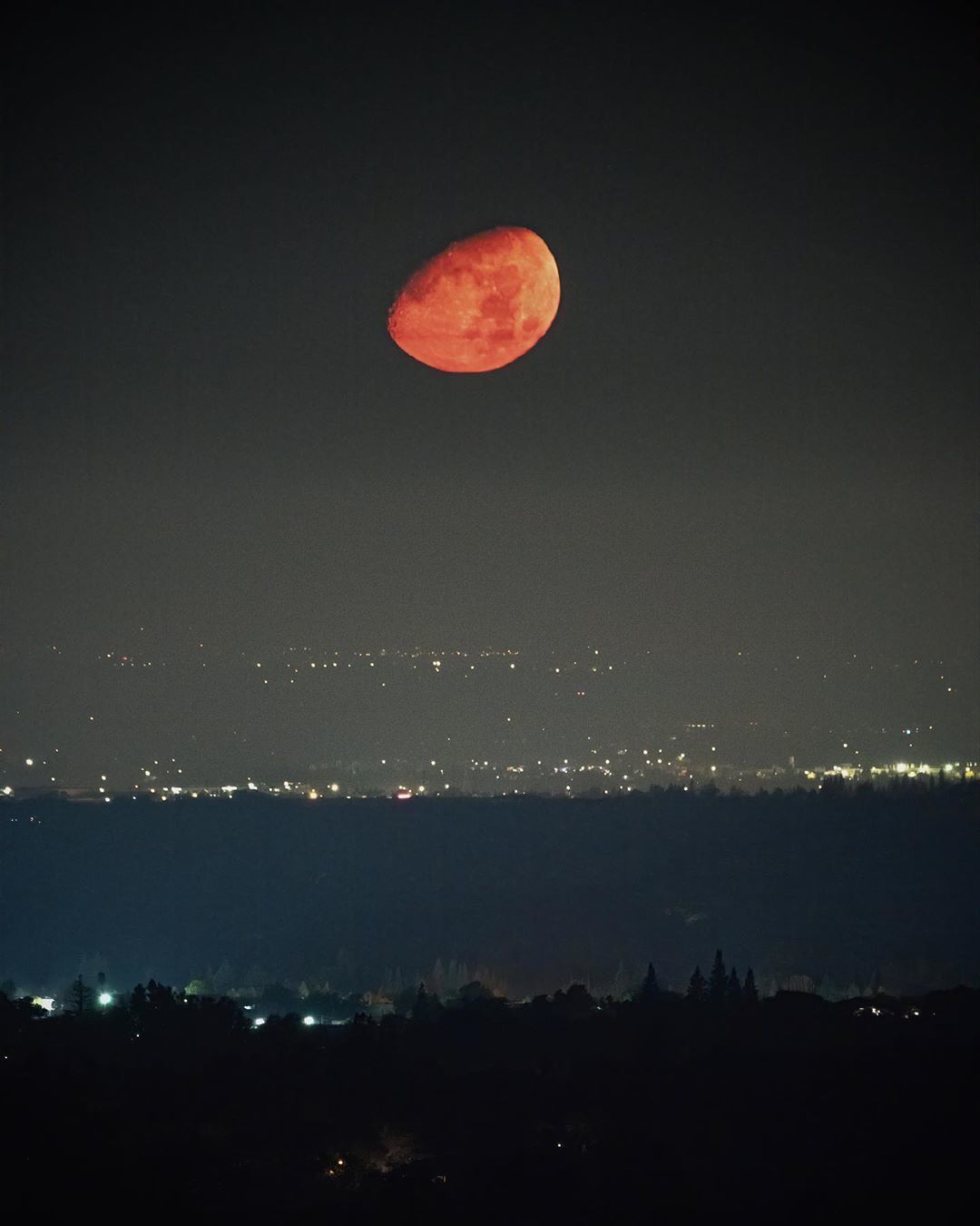 A red moon hangs over foothills ridge lines at night with city lights beyond