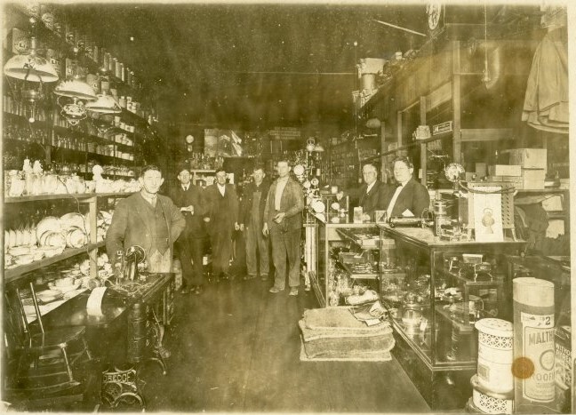 Worn photo depicting interior of Lukens Hardware Store, 1915, with owner Guy Lukens on the far right