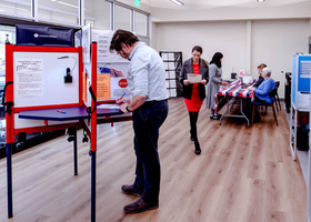 A man casts a ballot at a voting booth as other voters are checked in in the background of a voting center.