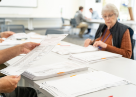  A woman seated at a table reviews stacks of voted ballots on a table in front of her.