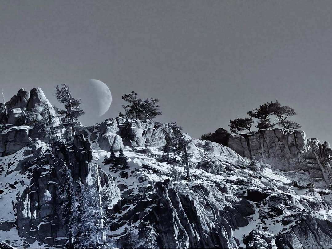 Black and white photograph of mountains covered in snow and some trees, half moon in background behind mountians. 