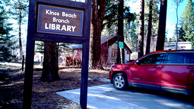 A close-up of the Kings Beach branch library sign in North Lake Tahoe in front of trees and next to a red car. 