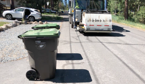 A green waste trash can on an asphalt street with a truck approaching in the background. 