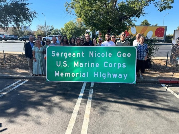  A group of people standing behind a green metal highway sign that reads, Sergeant Nicole Gee U.S. Marine Corps Memorial Highway
