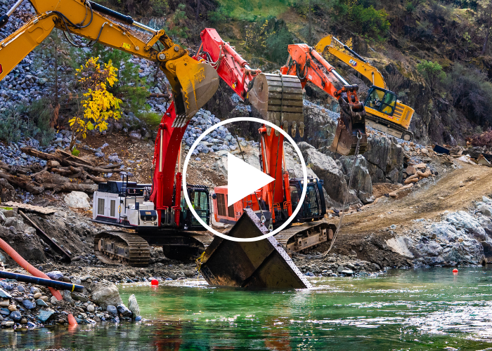 An excavator removing a large piece of metal from the river, with a play arrow over the photo