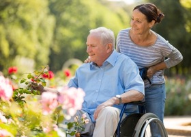 Woman caregiver pushing an elderly man in a wheelchair, outside in the sun near some flowers