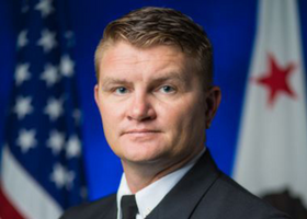 Jim Hudson poses in a navy blue suit before the United States and California flags