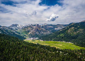 Aerial image of Olympic Valley on a cloudy day with vast green trees, open green fields and snow-capped mountains.