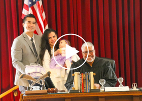 Woman holding a baby standing with a man behind a judge, all smiling in a courtroom