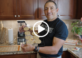 A man poses in a kitchen with a rack of cookies with a play button icon layered on top