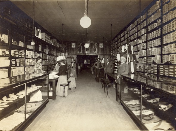Antique photograph of men and women in period attire shopping in a store lined floor to ceiling with shelves and goods