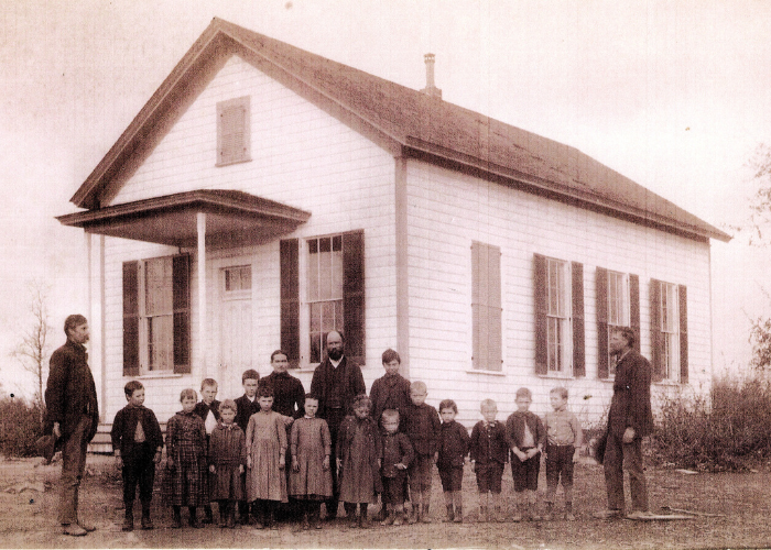 Students and adults standing in front of a one-room schoolhouse