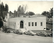 A historic photo of a white, one-story building with period automobiles parked on tihe street in front