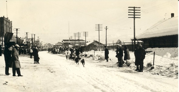 A dog sledder races between spectators who are lined up in front of buildings