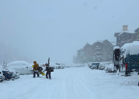 White-out conditions at the Village of Palisades Tahoe with two skiers carrying gear in the parking lot.