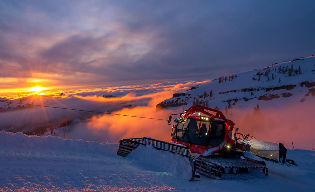 A red snow plow grooms snow on a ski run along a mountain side. The morning sun beams across the morning clouds.