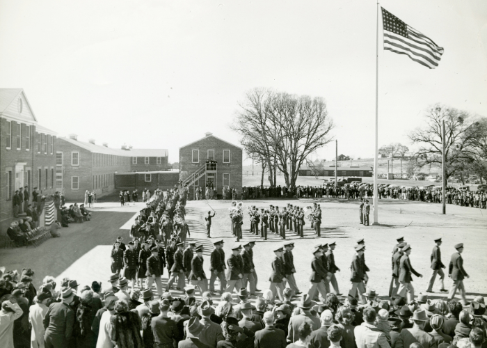 Military officers march in formation before onlookers in front of a military hospital building