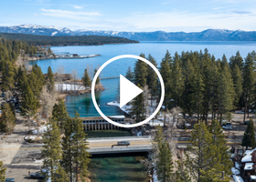 An aerial image of a truck going over Fanny Bridge with Lake Tahoe’s blue water and surrounding mountains in the background with a video play button