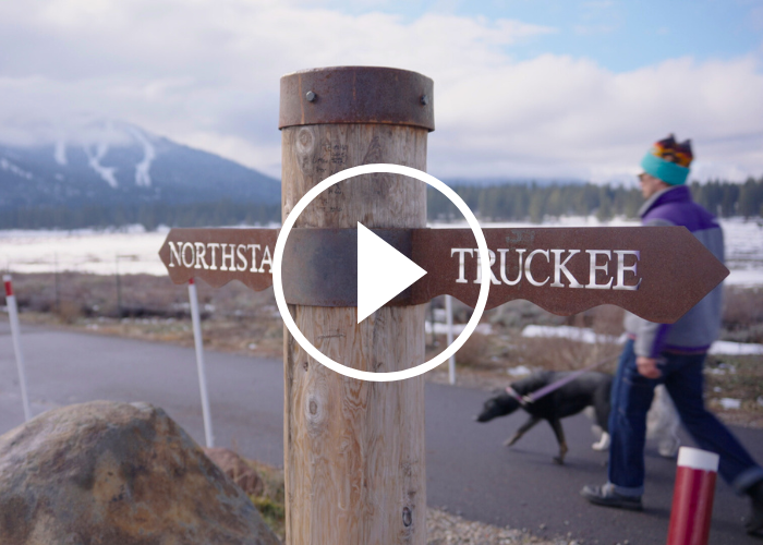A wooden post with metal signs designating the Town of Truckee to the right and Northstar to the left with a dog walker in the background.