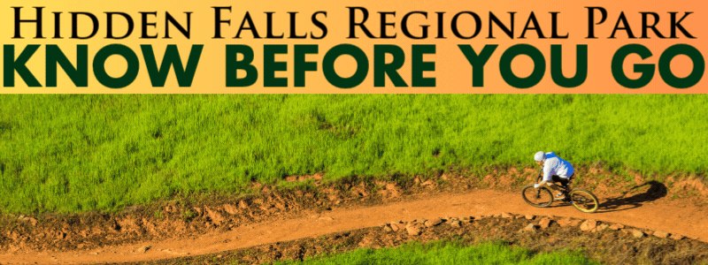 Mountain biker in a white shirt on a single track dirt trail surrounded by green fields. Text reads Hidden Falls Regional Park know before you go