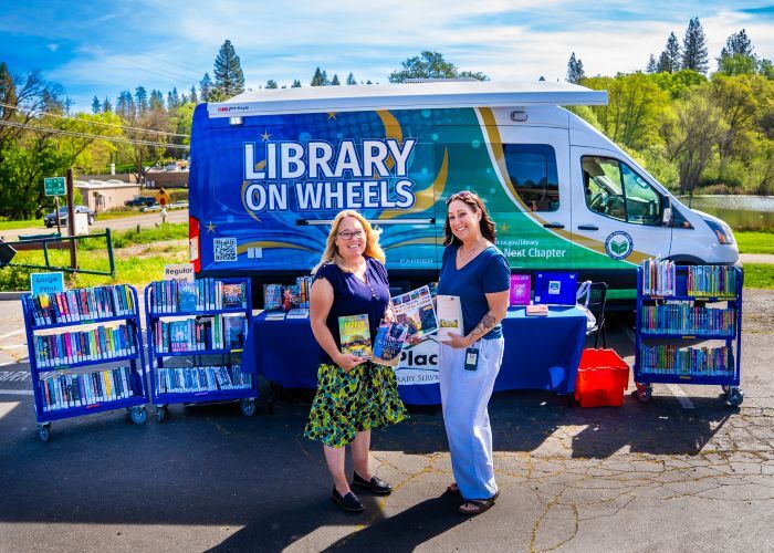 Two librarians pose holding books in front of the new library on wheels and a book display at Meadow Vista Park