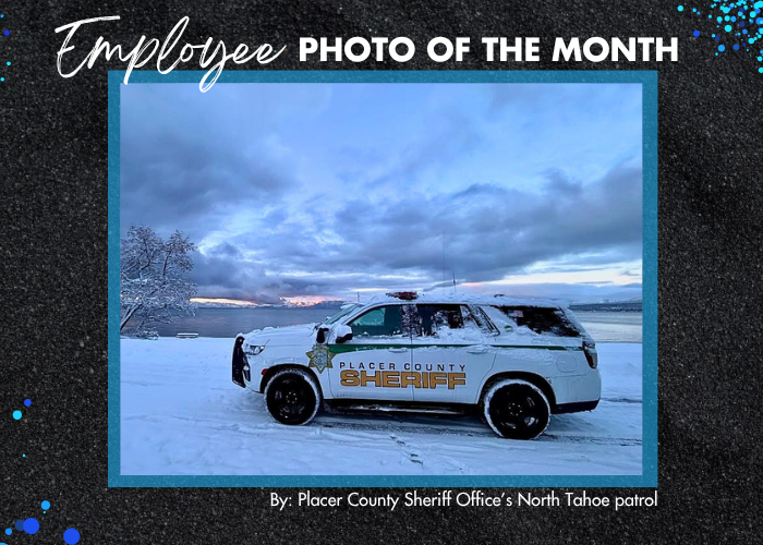 A white SUV covered in snow with yellow Placer Sheriff lettering on the side in front of Lake Tahoe with a pink sunrise in the distance.