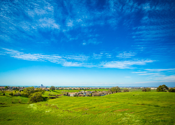 A green field in front of rows of homes under a large blue sky with wispy white clouds.