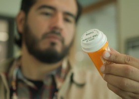 Man holding an orange prescription pill bottle and reading the label indoors