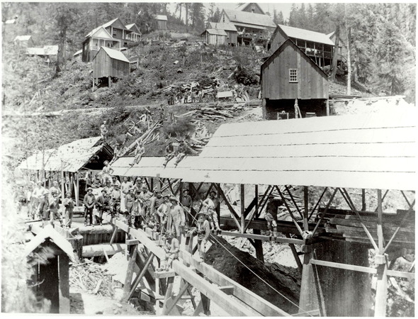 Men stand in a line in front of a mine with small houses built on stilts up a steep slope