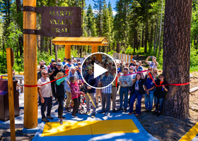 A group of people cut a red ribbon with large gold scissors at the Martis Valley Trail entrance on Northstar Resort property.