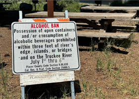 A white and orange sign with black and red text stating the county’s alcohol ban on the Truckee River from July 1-6 in front of picnic benches