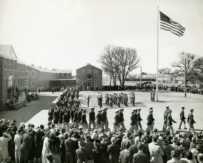 Dozens of soldiers in dress uniform march through parade grounds with a crowd looking on at the DeWitt General Hospital opening ceremony