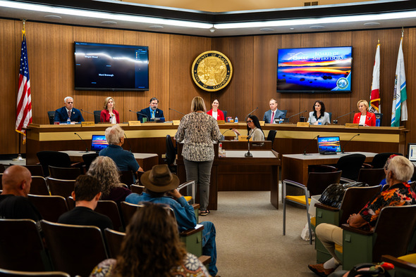 Board of Supervisors members listen as a woman speaks from a podium in a wood-paneled chamber with flags and screens.
