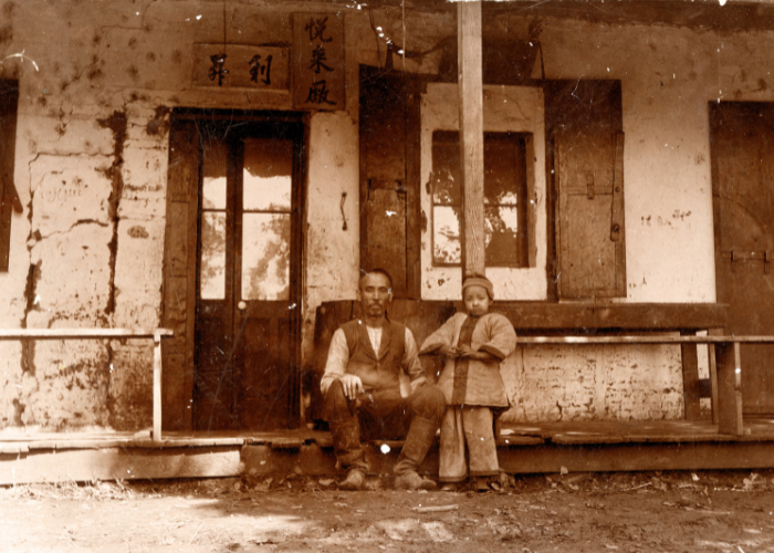A small child dressed in traditional Chinese clothing stands next to a man sitting on porch of a Chinese storefront