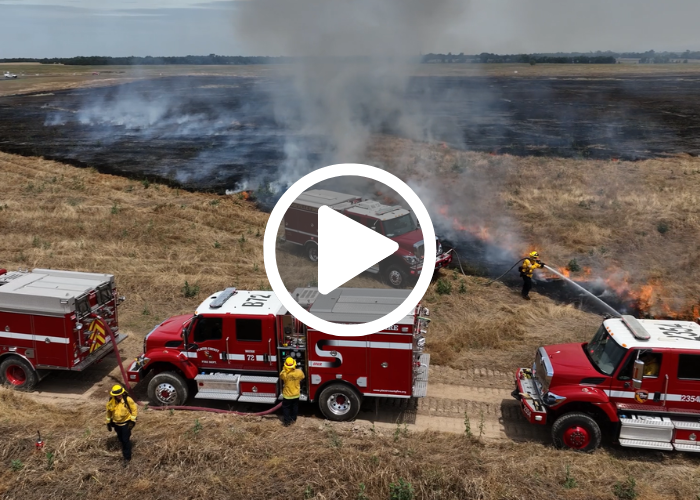 4 red Type 2 wildland fire engines with multiple firefighters spraying water on a fire in a yellowed field with smoke and a video play button overlay.