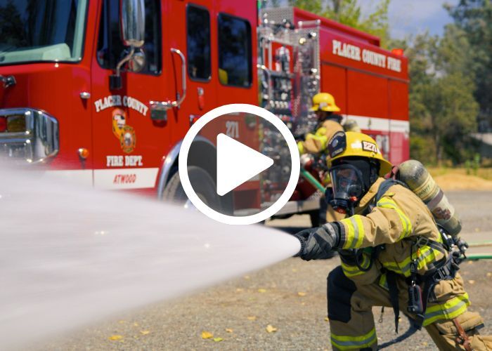 Firefighter sprays water from a fire hose while in a stance in front of a fire engine with another firefighter working by the truck.